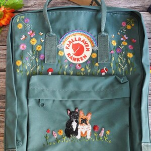 May include: Teal backpack with embroidered floral and butterfly details. Features a rainbow patch with the Fjallraven Kanken logo. The front pocket showcases embroidered French Bulldogs among flowers. Includes top handles and adjustable straps.