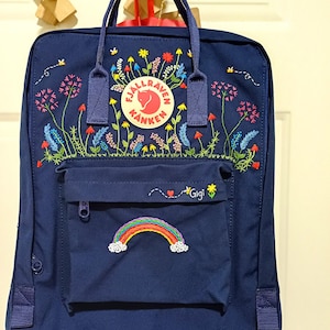 Puede incluir: Una mochila azul marino con diseños florales y arcoíris bordados. La mochila tiene un bolsillo frontal con cremallera y el nombre "Gigi" bordado sobre un arcoíris. Tiene un logotipo redondo con "Fjallraven Kanken".