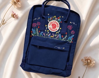 May include: Navy blue backpack with embroidered floral design. The backpack features a front pocket with a zipper and a top handle. The embroidered flowers include pink, blue, and yellow colors. The backpack has the brand logo in the center.