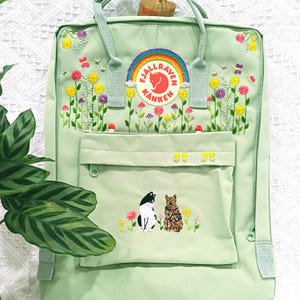 May include: A light green backpack with embroidered flowers, butterflies, and bees. The backpack features a rainbow logo and a front pocket with embroidered cats. The bag has a top handle and adjustable straps.