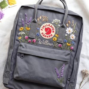 Puede incluir: Mochila gris con bordados de flores silvestres multicolores. La mochila presenta el logotipo de Fjallraven Kanken y el nombre "Willow" bordado. Hilos de bordar verdes, amarillos y morados son visibles en el fondo.
