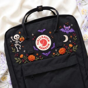 Puede incluir: Una mochila negra Fjällräven Kånken con bordados detallados de un esqueleto sosteniendo una calabaza, un murciélago morado sonriente, una luna creciente, flores naranjas y pequeños murciélagos morados. El logo circular está centrado.