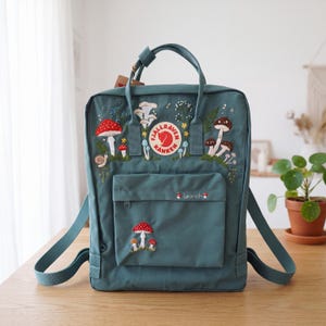 May include: A teal Fjällräven Kånken backpack featuring embroidered mushroom, snail, and floral motifs. The front pocket displays the name "Leanah" in white script with a small red mushroom. It includes top handles and adjustable shoulder straps.