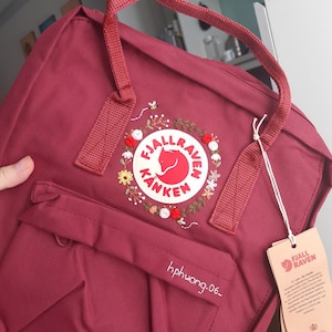 Könnte beinhalten: Ein burgunderfarbener Fjällräven Kånken Rucksack mit einem weißen gestickten Logo und floralen Details. Der Rucksack hat eine kleine Tasche auf der Vorderseite mit dem gestickten Text "hphwong.06_" .