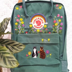 May include: A teal backpack with embroidered flowers, butterflies, and a rainbow patch. The front pocket features embroidered cats and the name "Pia". The backpack has a top handle and adjustable straps.
