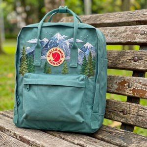 Könnte beinhalten: Türkisfarbener Rucksack mit gesticktem Berg- und Kieferndesign. Mit einer Vordertasche, einem oberen Griff und dem Fjallraven Kanken-Logo. Der Rucksack liegt auf einer Holzbank.