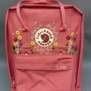 Puede incluir: Una mochila rosa con el logotipo bordado de Fjällräven Kånken, rodeado de flores silvestres coloridas, pequeñas setas y abejas. El bolsillo delantero muestra el nombre bordado Suktika flanqueado por dos pequeños iconos de arcoíris.