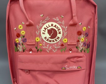 May include: A pink backpack featuring an embroidered Fjällräven Kånken logo, surrounded by colorful wildflowers, small mushrooms, and bees. The front pocket displays the embroidered name Suktika flanked by two small rainbow icons.