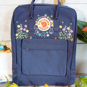 May include: A navy blue Fjällräven Kånken backpack featuring a circular logo patch and a colorful embroidered design of wildflowers, small mushrooms, and whimsical stick figures surrounding the brand name and fox emblem on the front pocket.