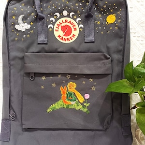 May include: A dark gray backpack with embroidered celestial and Little Prince-themed designs. The front pocket features an embroidered scene of the Little Prince and a fox. The top has embroidered stars, moons, and planets.