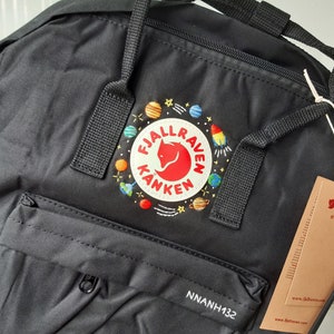 May include: Black backpack with a front zippered pocket and top carry handles. The backpack features a circular patch with a red fox logo and colorful space-themed embroidery. The text "FJALLRAVEN KANKEN" is on the patch. The tag reads "www.fjallraven.com".