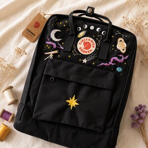 Puede incluir: Mochila negra con diseños celestiales bordados, incluyendo una luna creciente, planetas y estrellas. Cuenta con un bolsillo frontal con una estrella amarilla y la etiqueta de la marca "Kanken Original". Incluye asa superior y correas ajustables.