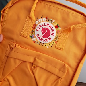 Puede incluir: Mochila de color amarillo mostaza con un bolsillo frontal y asa superior. La mochila presenta el logotipo "Fjallraven Kanken" en un círculo rojo con un emblema de zorro, rodeado de flores bordadas y notas musicales.