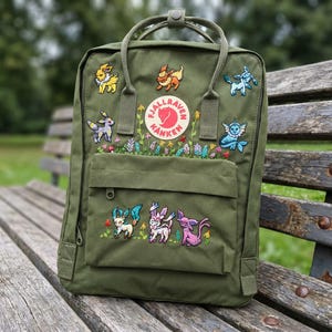 May include: Olive green backpack with embroidered Pokémon characters and floral accents. Features a front pocket and top handle. The backpack has a round logo patch with the text "Fjallraven Kanken".