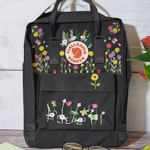 May include: Black Fjällräven Kånken backpack with embroidered floral designs. The backpack has a small front pocket and adjustable straps.