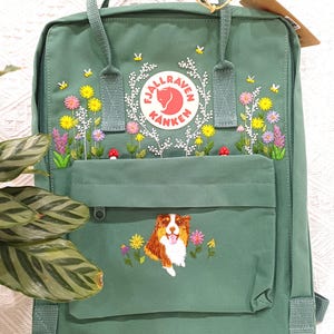 Peut inclure: Un sac à dos vert avec des broderies florales et un motif de chien. Le sac à dos comporte une poche avant avec une broderie de chien et un compartiment principal avec un motif floral autour du logo. Le sac a des poignées supérieures et des bretelles réglables.