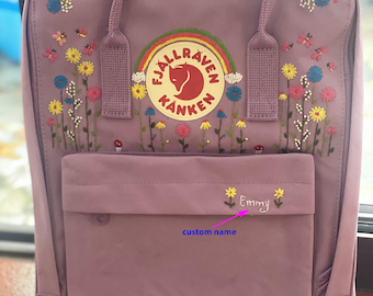 May include: A lavender backpack with embroidered flowers, butterflies, and the text "Emmy". The backpack features a front pocket and the Fjallraven Kanken logo with a rainbow. The bag has two top handles and adjustable shoulder straps.