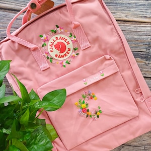 Puede incluir: Una mochila rosa Fjällräven Kånken con bordado floral personalizado alrededor del logotipo circular y en el bolsillo delantero. El bordado incluye flores coloridas y el nombre Clodagh cosido en hilo rosa en la solapa del bolsillo.