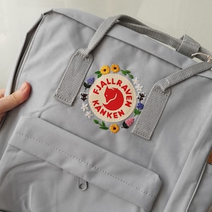 May include: A light gray backpack with a front pocket and top handles. The backpack features a circular patch with the text "FJALLRAVEN KANKEN" and a fox logo, surrounded by embroidered flowers in yellow, white, and purple.