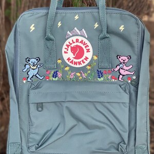 May include: A slate blue backpack featuring an embroidered Fjällräven Kånken logo, two dancing bears, colorful wildflowers, and small yellow lightning bolts. The bag includes a front zippered pocket, two top handles, and adjustable shoulder straps.