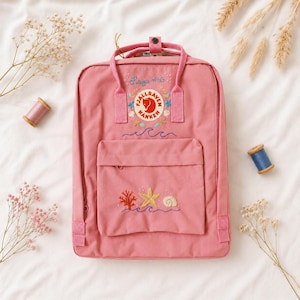 May include: A pink backpack with embroidered ocean-themed designs, including waves, a starfish, and coral. The backpack features the text "Raiya x HG" and the Fjallraven Kanken logo. It has a top handle and front pocket.