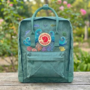 May include: Teal backpack with embroidered ocean design, featuring dolphins, waves, and coral. Includes a front pocket and top handle. The backpack has a circular logo with text.