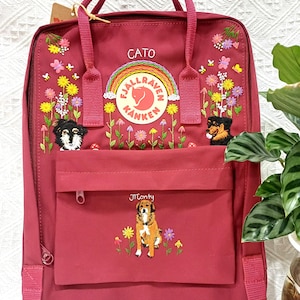 May include: A deep red backpack featuring embroidered floral patterns, butterflies, and three dogs. The Fjällräven Kånken logo is centered with a rainbow arch above it. The names CATO and Monty are embroidered in white thread above the dog portraits.