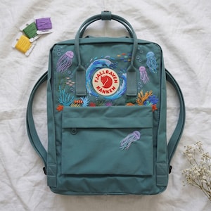 Könnte beinhalten: Ein blaugrüner Rucksack mit einer gestickten Unterwasserszene, die eine zentrale Welle, Quallen, Korallen und kleine Fische zeigt. Das kreisförmige Fjällräven Kånken-Logo befindet sich mittig über einer aufgesetzten Fronttasche.