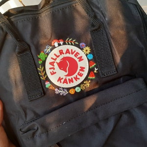 May include: Black backpack with embroidered floral and fruit details around the Fjallraven Kanken logo. The embroidery includes flowers, mushrooms, cacti, watermelon slices, and a bee. The backpack has black straps and a top handle.
