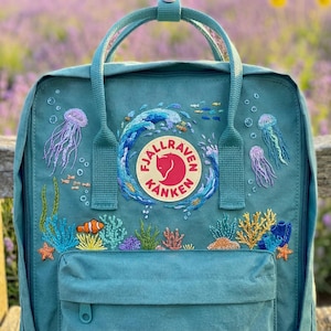 May include: Teal backpack with embroidered ocean design. Features a central logo and detailed embroidery of jellyfish, coral, fish, and a wave. Includes a front pocket and top handle. The background is a blurred view of purple flowers.