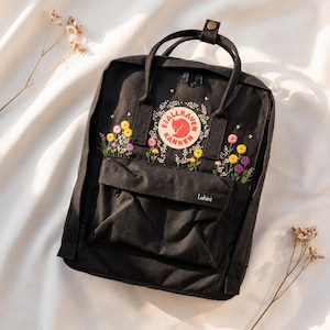 Puede incluir: Mochila negra con diseño floral bordado en amarillo, morado y rosa. La mochila presenta un logotipo redondo con un zorro rojo y el texto "Fjallraven Kanken". El nombre "Lahini" está bordado en el bolsillo delantero.