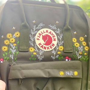 May include: A green Fjällräven Kånken backpack with embroidered flowers, bees, and mushrooms. The logo is visible on the front of the bag, and the name "Nati" is embroidered below the pocket.