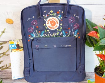 May include: Navy blue backpack with embroidered floral design. The backpack features a front pocket with heart and floral embroidery. The backpack has a top handle and adjustable shoulder straps. The backpack has the Fjallraven Kanken logo.