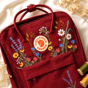 Op de afbeelding: Een bordeauxrode rugzak met geborduurde wilde bloemen, waaronder madeliefjes, lavendel en korenbloemen. De rugzak heeft een voorvak, een handvat aan de bovenkant en het Fjallraven Kanken-logopatch. De borduursels geven een ambachtelijk, artistiek tintje.