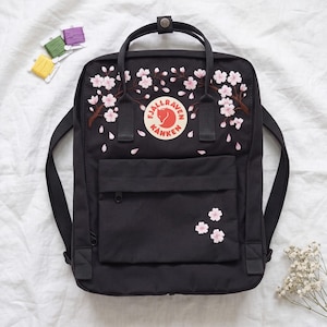 Puede incluir: Una mochila negra Fjällräven Kånken con ramas de cerezo en flor bordadas en rosa y pétalos cayendo sobre la parte superior y el bolsillo frontal. El logotipo circular está centrado, e incluye dos asas superiores y correas para los hombros.