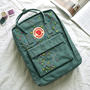 Op de afbeelding: Een teal rugzak met kleurrijke geborduurde bloemmotieven. De rugzak heeft een voorvak en een handvat aan de bovenkant. De tekst "HI" is geborduurd in tekstballonnen. Een ronde patch met een rood berenlogo is zichtbaar.
