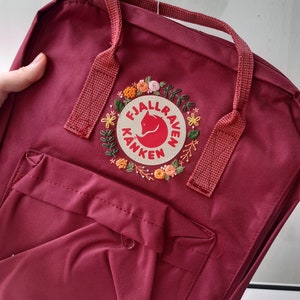May include: A burgundy backpack with a floral embroidered patch featuring a red fox logo. The backpack has a front pocket, top handles, and a zipper closure. The bag is made of a durable fabric.
