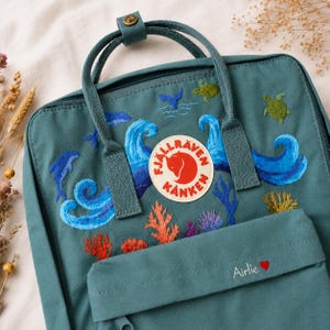 May include: Teal backpack with embroidered ocean scene, including waves, dolphins, whales, and coral. Features the Fjallraven Kanken logo and the name "Airlie" with a heart. The bag has a top handle and adjustable straps.