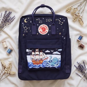 May include: A dark blue Fjällräven Kånken backpack featuring embroidered constellations and a crescent moon on the upper section. The front pocket displays an embroidered scene of a sailing ship on blue waves with a light blue octopus.
