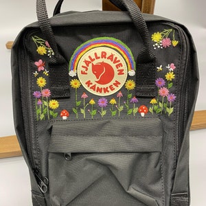 May include: A charcoal gray backpack featuring a circular Fjällräven Kånken logo patch. The bag is decorated with embroidered colorful wildflowers, small red mushrooms, a rainbow arching over the logo, and tiny bees on the front panel.