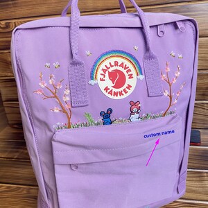 May include: A light purple backpack featuring an embroidered rainbow, a fox logo, and the text Fjällräven Kånken. The design includes cherry blossom trees, small bees, and two small characters. A front pocket displays the text custom name.