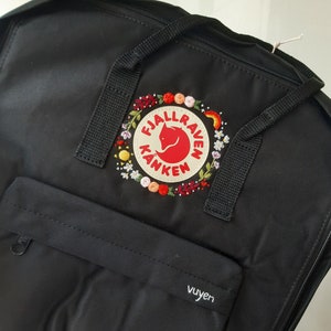 May include: Black Fjällräven Kånken backpack with a floral embroidered patch featuring the brand logo and the words "Fjällräven Kånken".
