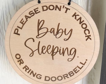 Könnte beinhalten: Ein rundes Schild aus Holz mit dem Text "Please Don't Knock Baby Sleeping Or Ring Doorbell." Das Schild hängt an einer Tür neben einer schwarzen Türklingelkamera.
