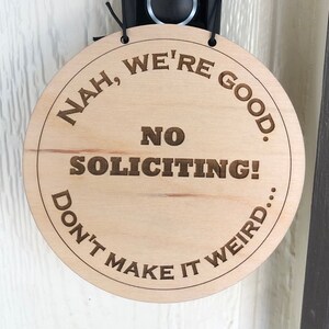 May include: A round wooden sign with the text "Nah, we're good. No soliciting! Don't make it weird..."  The sign is hanging on a door next to a black doorbell camera.