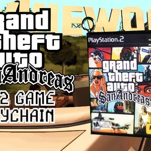 May include: A Grand Theft Auto: San Andreas PS2 game keychain. The keychain displays the game's cover art, including the logo and character images. The PlayStation 2 logo is visible. The background shows the Hollywood sign.