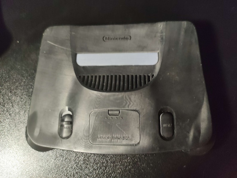 Op de afbeelding: Een zwarte Nintendo 64 console met een geheugenuitbreidingssleuf en een aan/uit-knop en een resetknop.