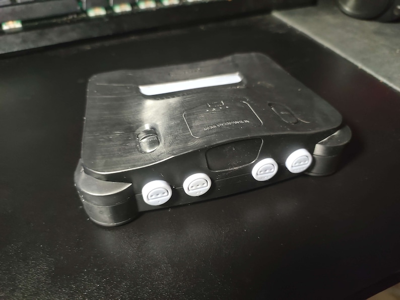 Op de afbeelding: Een zwarte Nintendo 64 console met grijze controllerknoppen. De console heeft een getextureerd oppervlak en een klein label met de tekst "MADE IN JAPAN".