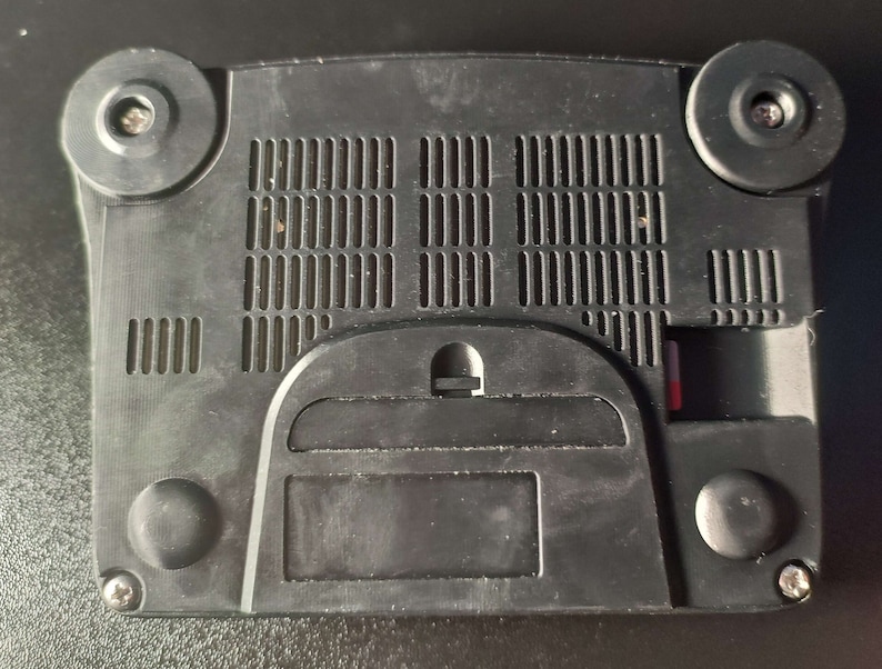 Op de afbeelding: Zwarte Nintendo 64 console met een ventilatie rooster aan de achterkant. De console heeft een slot voor een game cartridge en een klein klepje aan de achterkant.