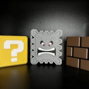 May include: Three-dimensional models of iconic video game elements. A yellow question mark block, a gray Thwomp with an angry face, and a brown brick wall are displayed against a dark background.