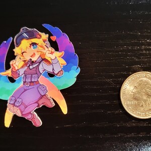 May include: A colorful sticker featuring a cartoon character with yellow hair, wearing a purple and grey outfit, and a rainbow-colored wing behind her. The sticker is about the size of a US quarter.